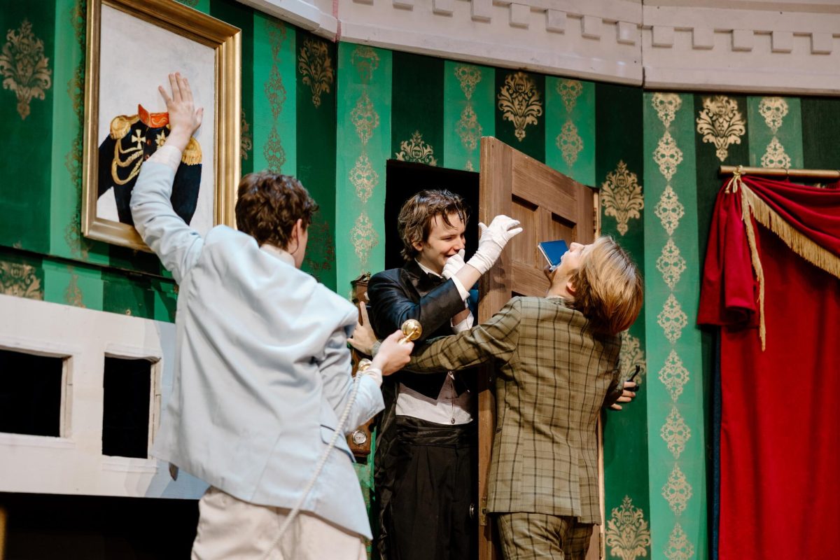 During this moment in The Play That Goes Wrong, we have Andrew Stoffers, Austin Wessel and Liam Dale attempting to hold the set together and keep it all from falling down as they carry on throughout the scene. Dale’s character, Robert, must take his checkbook from Wessel (as Dennis), while Stoffers’s character, Max, is holding up the portrait and waiting to hand the phone to Dale.