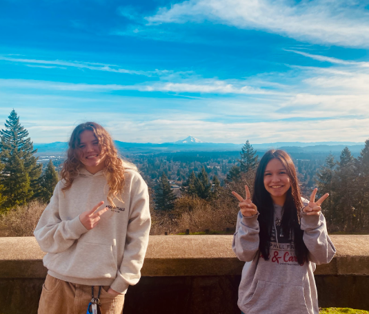 Two smiling girls posing in front of an outstretch of trees and a mountainside. Lyla R. and Esther M. were both on an Oregon City High School Geology Field Trip, held by Mr Nadeen the Geology Teacher at Oregon City High School, at Council Crest Park and were posing in front of Mt Hood.